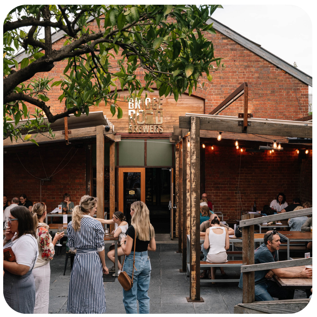 People enjoying outdoor seating at a brewery with brick building and wooden pergola.