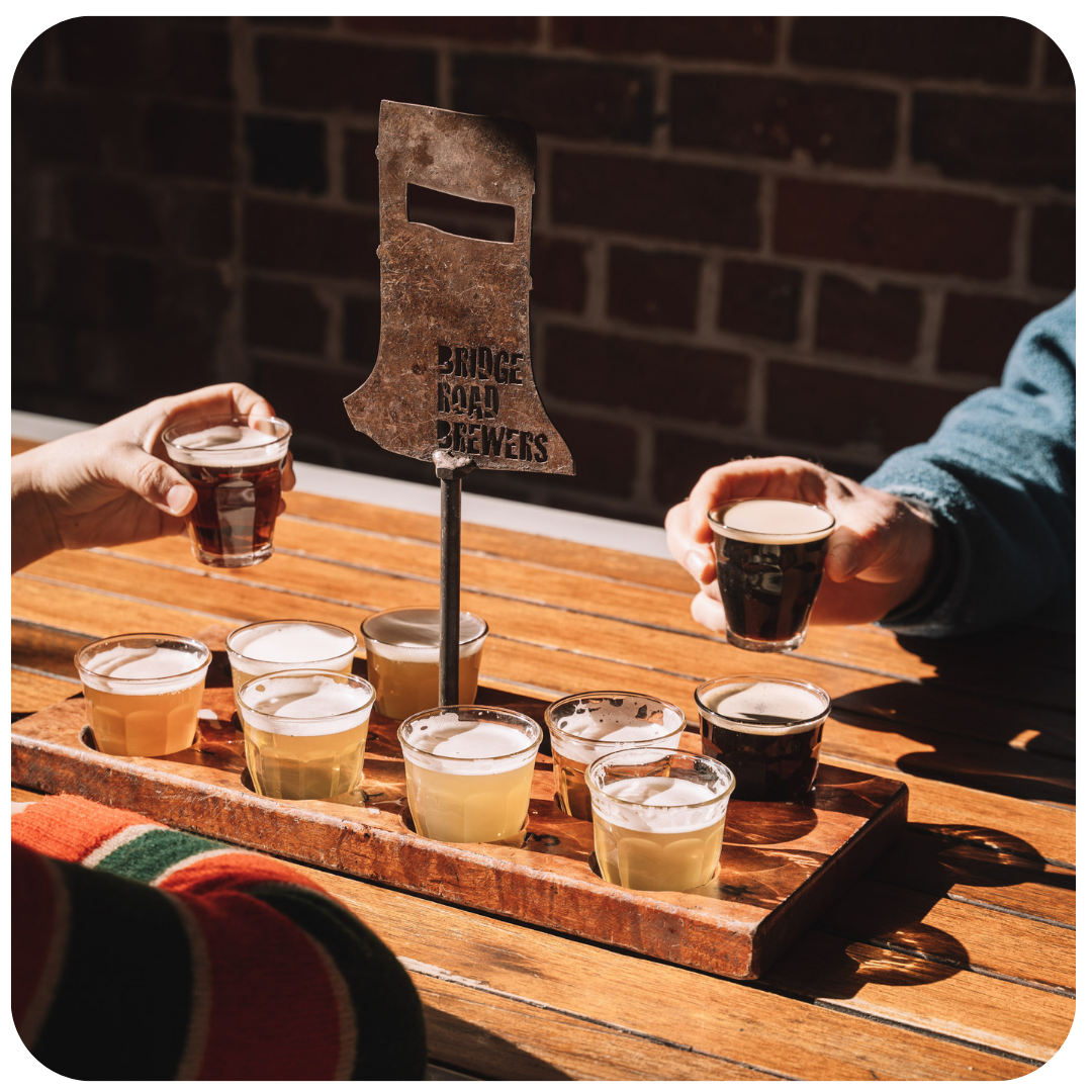 Two people holding glasses of beer with a wooden flight paddle displaying 'Bridge Road Brewers' against a brick wall.