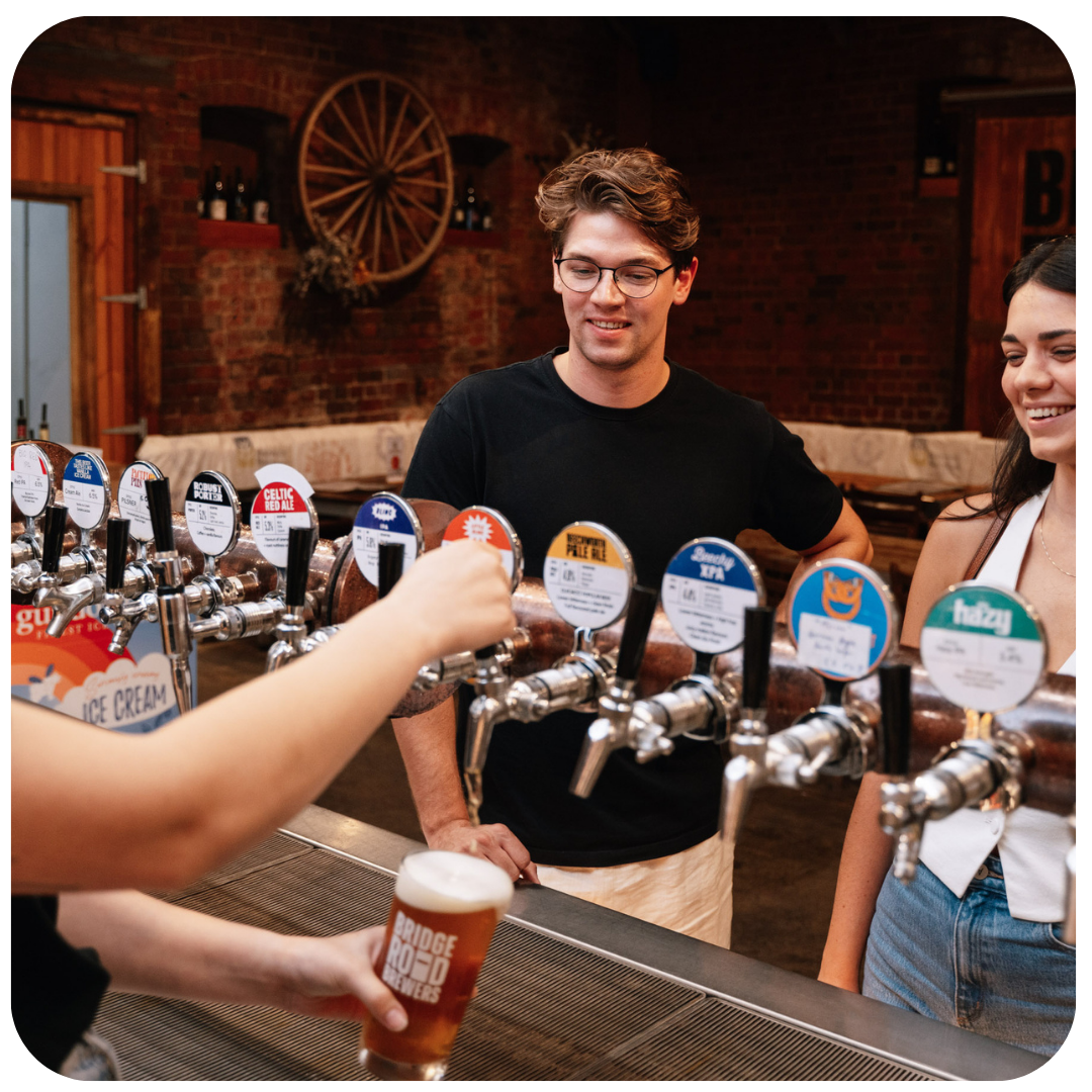 Person pouring a beer at a bar with a bartender and another person in the background.