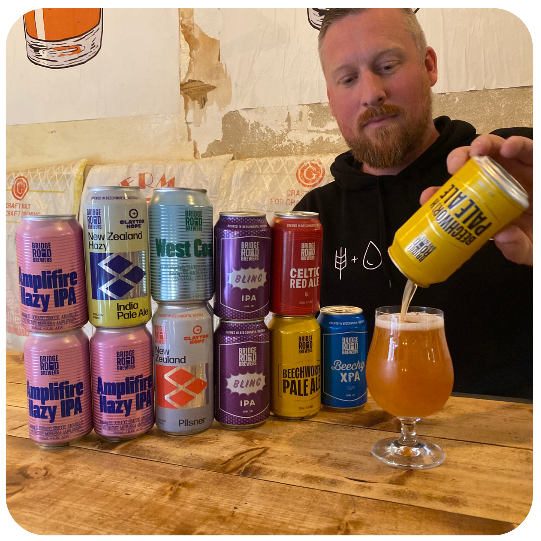Man pouring beer from a can into a glass with various beer cans on a wooden table.